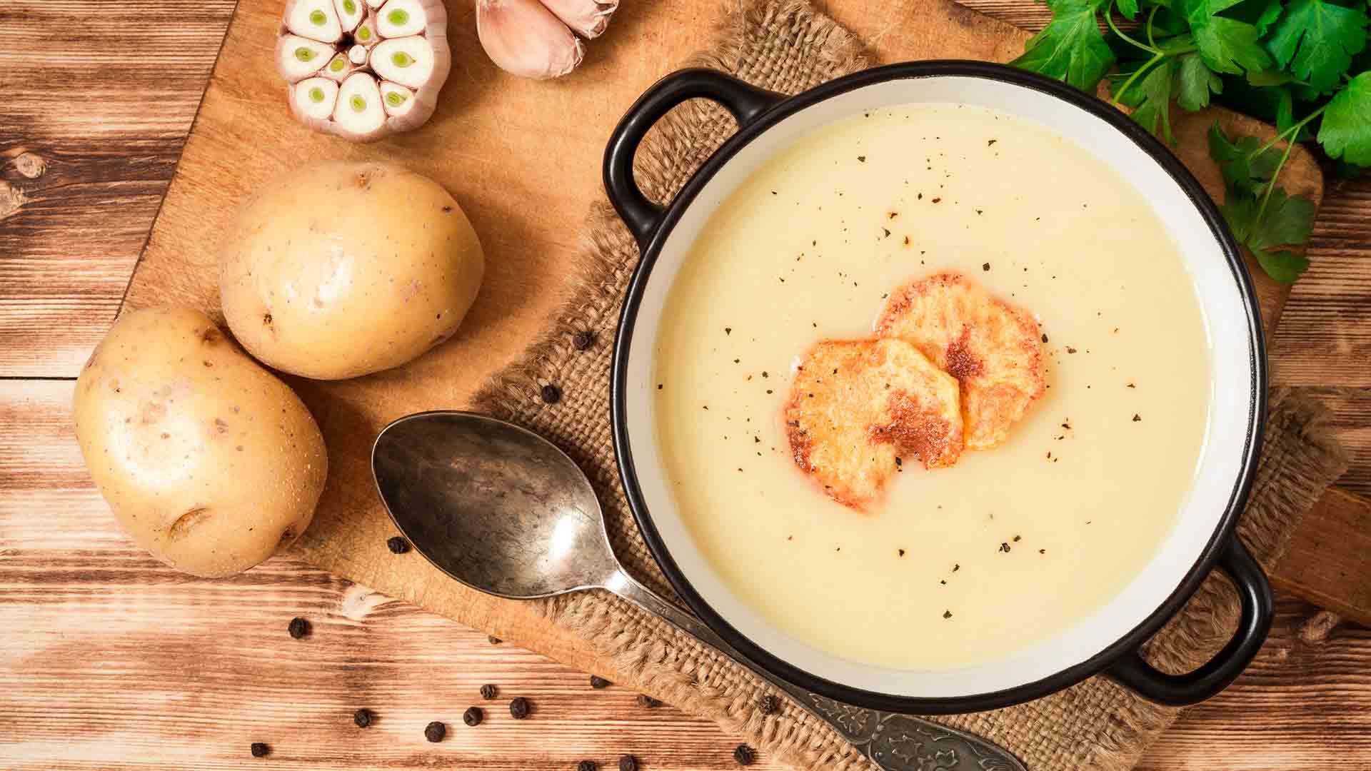 Potato Leaks Soup in a black bowl with potatoes and spoon near and it is placed in a wooden table
